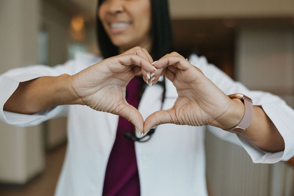 A person in a white coat holds hands in a heart shape. A stethoscope is visible around their neck.