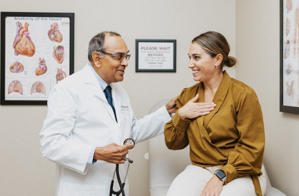 A doctor in a white coat and glasses converses with a smiling female patient in a mustard blouse, in a medical office with heart anatomy posters on the wall.