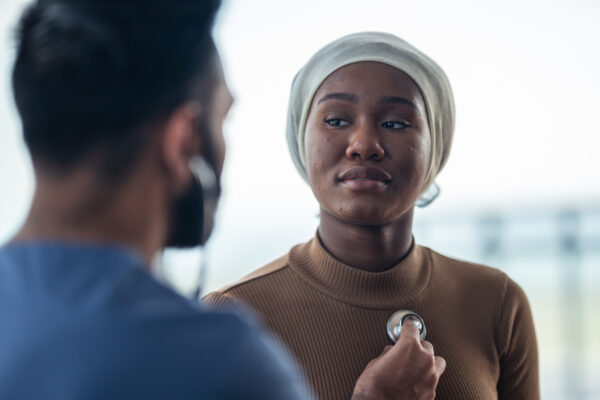 A healthcare professional listens to the chest of a patient using a stethoscope. The patient, wearing a light-colored headscarf and brown top, looks attentively at the healthcare professional. The background is softly blurred, focusing attention on them.