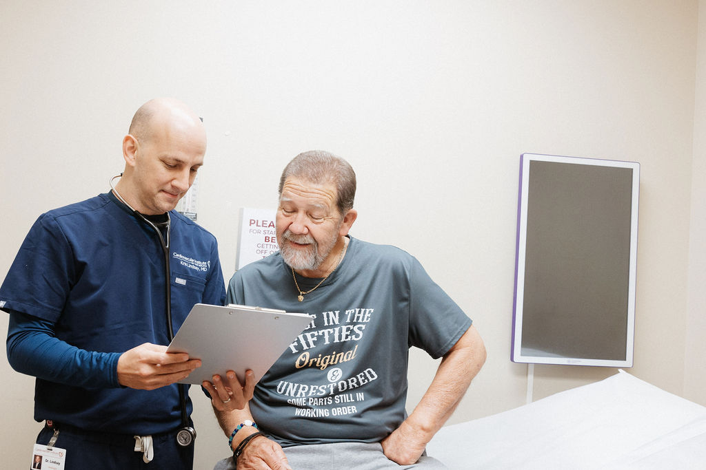 A healthcare professional in blue scrubs and a name badge shows a clipboard to an elderly man sitting on an examination table. The man is wearing a t-shirt and reading the clipboard attentively. The setting appears to be a medical examination room with a sterile environment.