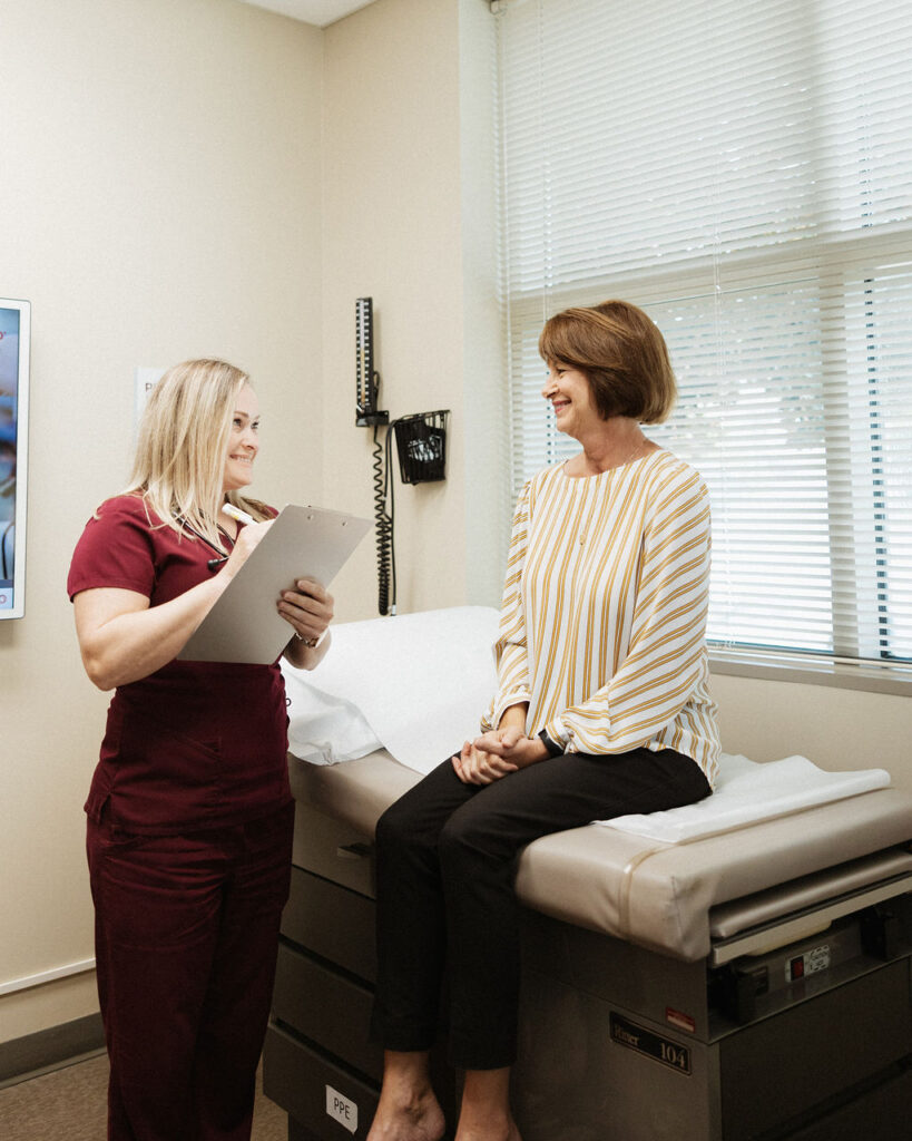 A cardiologist with a clipboard attentively listening to a smiling patient seated on an examination table in a medical office.