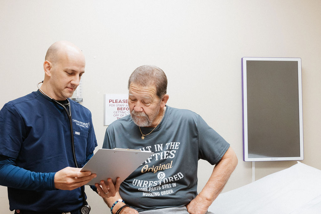 A healthcare professional in scrubs reviews a document on a clipboard with an attentive elderly male patient in a clinic room, discussing Venous Disease.