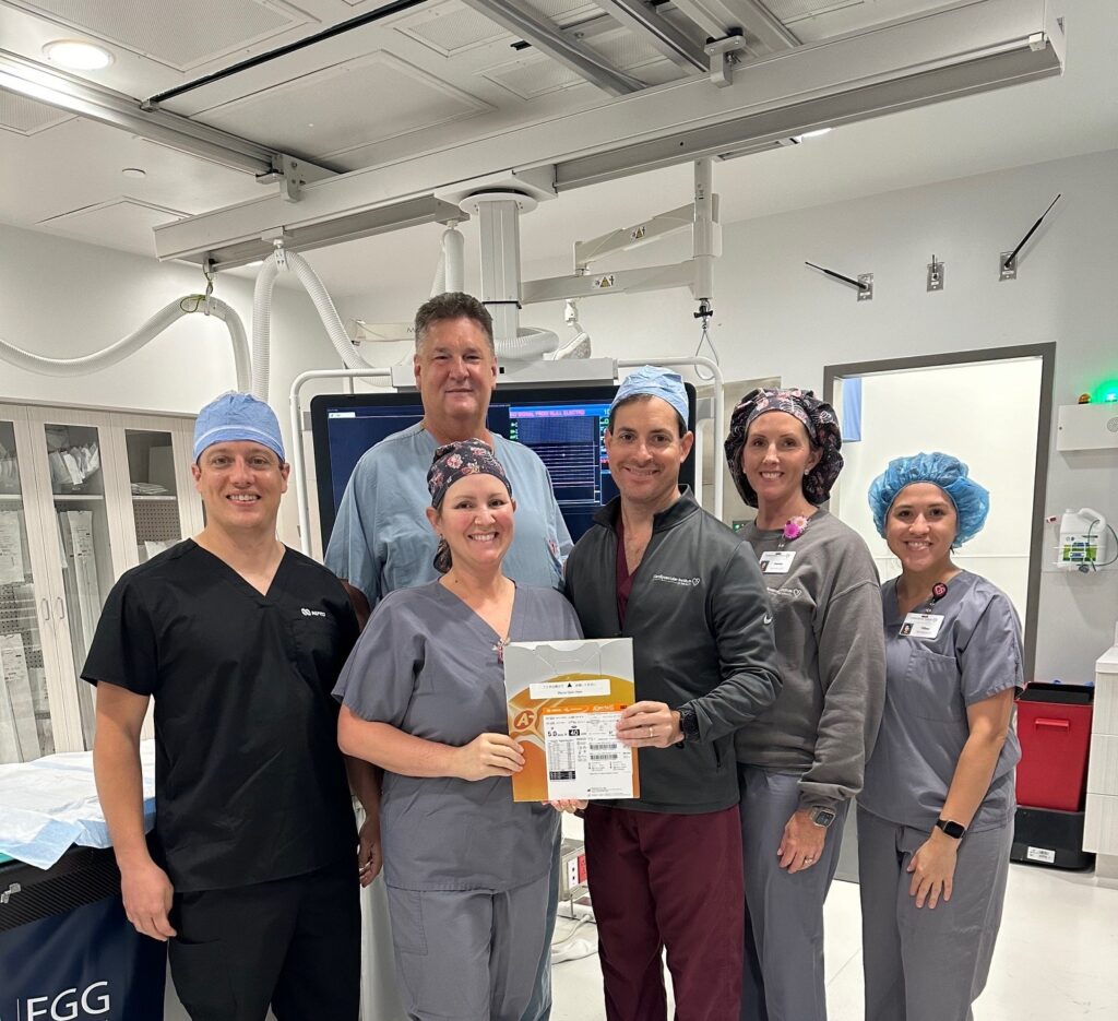 A group of six healthcare professionals, dressed in scrubs and surgical caps, smiling and standing together in a medical facility. One person is holding a certificate. Medical equipment and machinery are visible in the background.