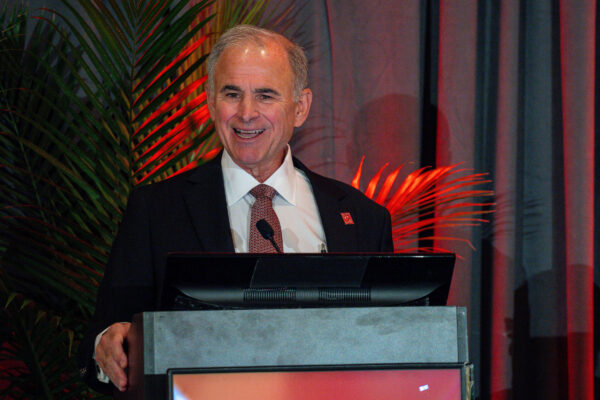 A man in a suit and tie is standing at a podium, speaking and smiling. He is framed by dark drapes and tropical plants illuminated with red and orange lighting. A computer monitor is on the podium in front of him.