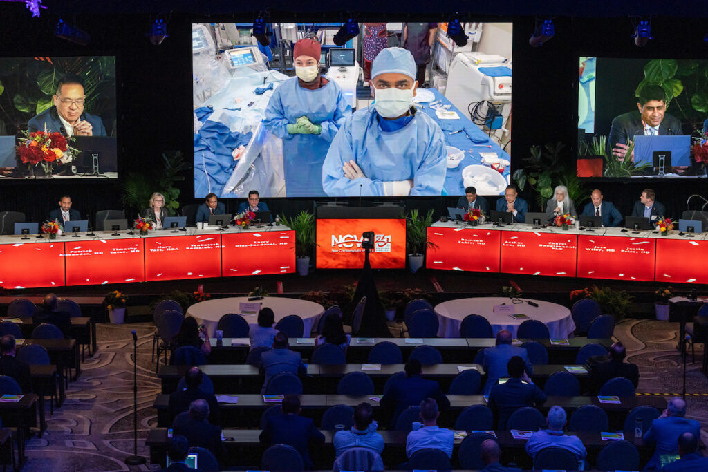 A conference room filled with attendees watching a large screen displaying a live surgery. Surgeons in blue scrubs, masks, and hair covers are shown on the screen. Panels of presenters sit at desks in front of the screen under red banners.