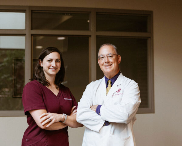Two medical professionals stand side by side inside a congestive heart failure clinic in Louisiana. The woman on the left is wearing maroon scrubs and has her arms crossed, while the man on the right is dressed in a white lab coat, also with arms crossed. Both are smiling and looking at the camera.