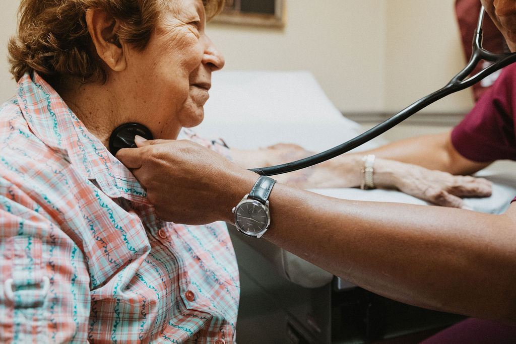 A heart specialist in Baton Rouge in scrubs uses a stethoscope to check the heartbeat of an elderly woman in a plaid shirt, who is sitting on an examination table in a clinic. The provider's arm, wearing a watch, is extended toward the woman's chest.