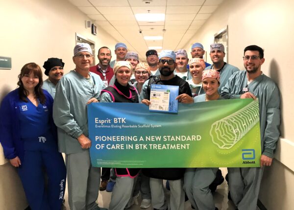 A group of healthcare professionals in scrubs stand in a hospital hallway, holding a green banner that reads, "Esprit BTK - Pioneering a new standard of care in BTK treatment" with the Abbott logo. They are smiling at the camera.