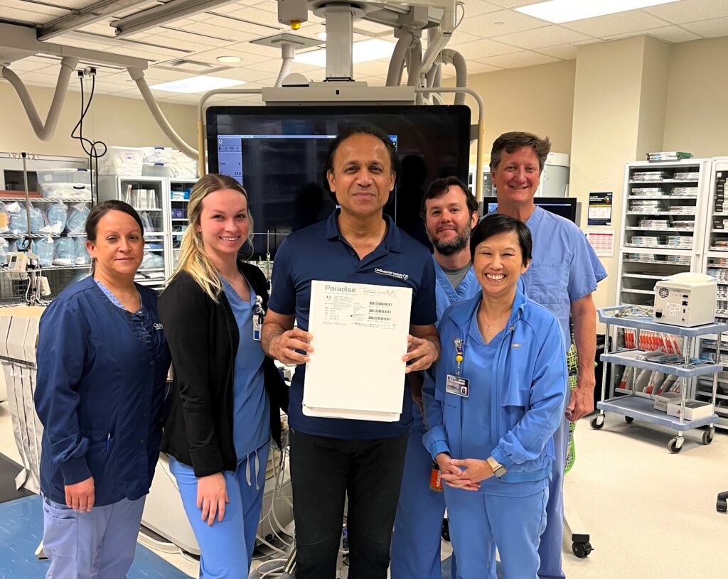 Six medical professionals stand in an operating room, dressed in scrubs. One person holds a box labeled "Paradise." They are surrounded by medical equipment and appear to be smiling at the camera. Kalyan Veerina of OGH Opelousas Cardiovascular Institute is noted for innovative treatments like renal denervation for hypertension.