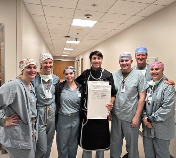 A group of seven healthcare professionals in scrubs and surgical caps pose for a photo in a hospital hallway. One individual in the center holds a protective lead apron and a sheet of paper. They all smile at the camera, suggesting a positive and supportive environment.