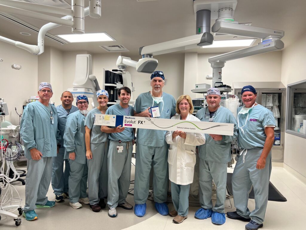 A group of medical professionals in scrubs and a lab coat stand together in an operating room, smiling at the camera. They are holding a large branded sign that reads "Evolut FX" and another item with the same branding. Medical equipment surrounds them.