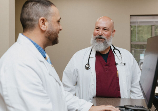 Two medical professionals in white lab coats converse in a clinic setting. One man, with a bald head and gray beard, stands with a stethoscope around his neck, smiling at his colleague who is facing him. The other man types on a computer.