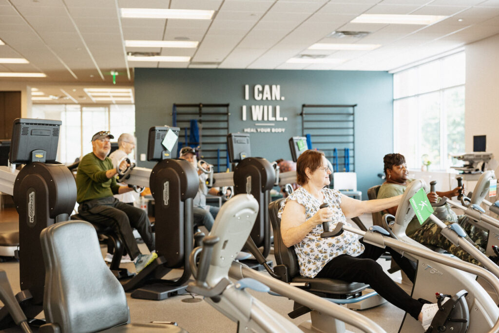 A group of people exercising on stationary bikes in a well-lit gym. In the background, a wall displays the motivational phrases "I CAN. I WILL. Heal Your Body." Various individuals, of different ages, appear focused and engaged in their workouts.