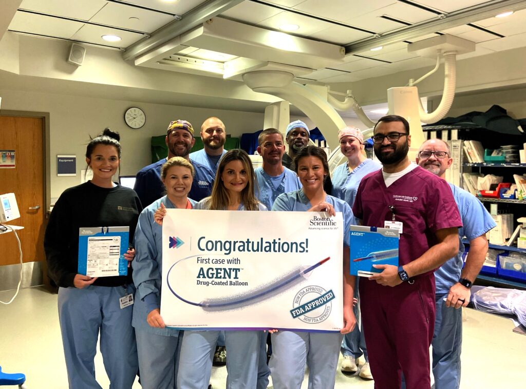 A group of healthcare professionals in scrubs are gathered in a medical room, smiling and holding a banner that reads, "Congratulations! First case with AGENT Drug-Coated Balloon." Some hold boxes labeled "AGENT" and official documents. The room is well-lit and contains medical equipment.