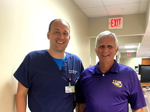 Two smiling men pose for a photo in a hallway at the Cardiovascular Institute of the South in Opelousas, Louisiana. The man on the left is wearing blue medical scrubs and has a name badge. The man on the right is wearing a purple polo shirt with a yellow logo. An exit sign is visible on the ceiling behind them.