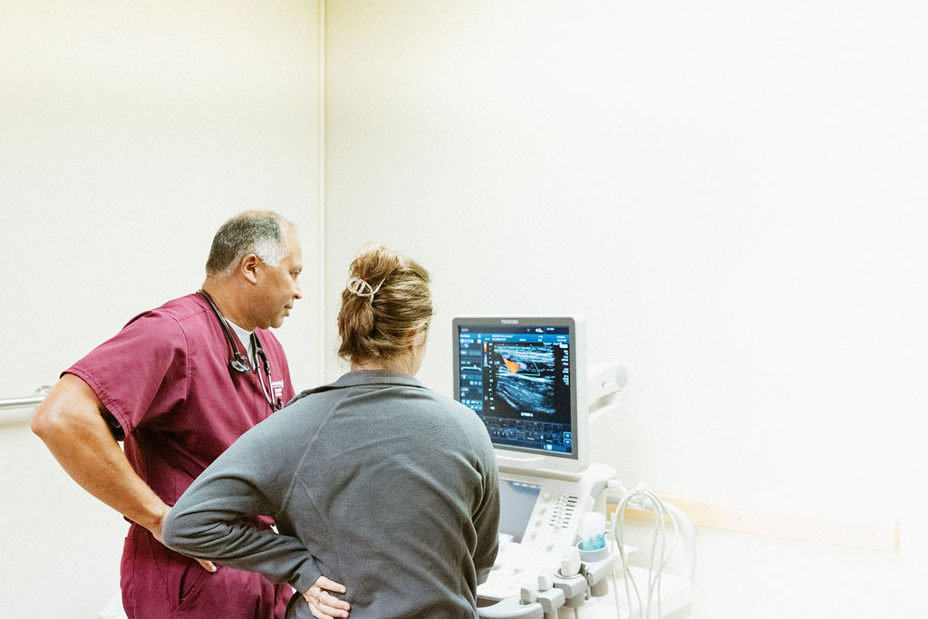 Two medical professionals, one in burgundy scrubs and the other in a gray jacket, stand in front of an ultrasound machine. They are intently looking at the monitor displaying ultrasound images in a well-lit examination room.