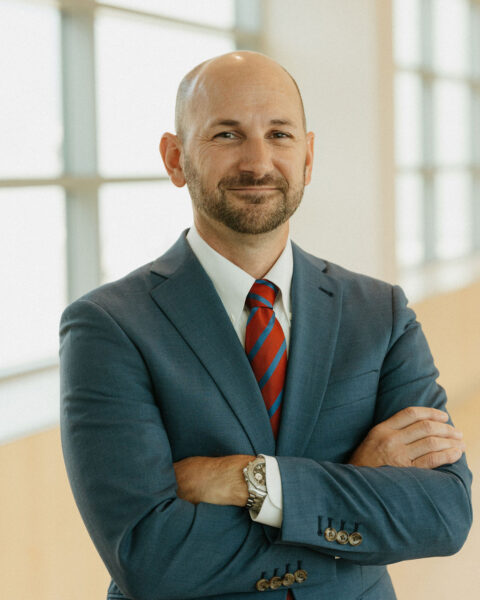 A bald man with a beard is standing with arms crossed, wearing a blue suit, white dress shirt, and red-striped tie. He is smiling slightly and posing in a well-lit room with large windows in the background.