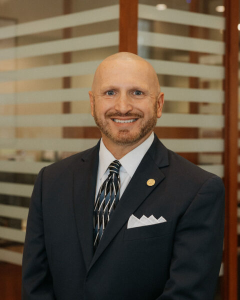 A bald man with a beard and a friendly smile is wearing a dark suit, a patterned tie, and a white pocket square. He stands in front of a background of glass panels with horizontal frosted stripes.