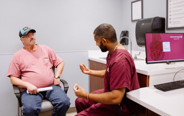 A man in a pink t-shirt and baseball cap sits on a chair, holding a piece of paper in his hand. He faces a healthcare professional in maroon scrubs, who is gesturing with his hands. They are in a medical office with a computer displaying a cardiac-related image.