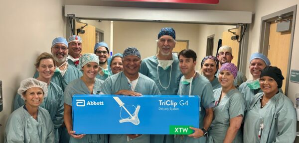 A group of healthcare professionals in scrubs and caps stand together, smiling at the camera. They are holding a large blue box labeled "Abbott TriClip G4 Delivery System" used for triclip valve repair. The scene is set in a hospital hallway, likely capturing a breakthrough moment for tricuspid valve treatment.