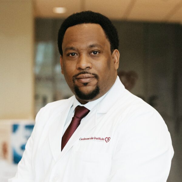 Dr. Marcus Cox, a cardiologist at Iberia Medical in New Iberia, Louisiana, stands indoors wearing a white lab coat with the "Cardiovascular Institute" logo on the chest and a red tie. He has a focused expression and a well-groomed beard. The softly blurred background suggests a professional setting.