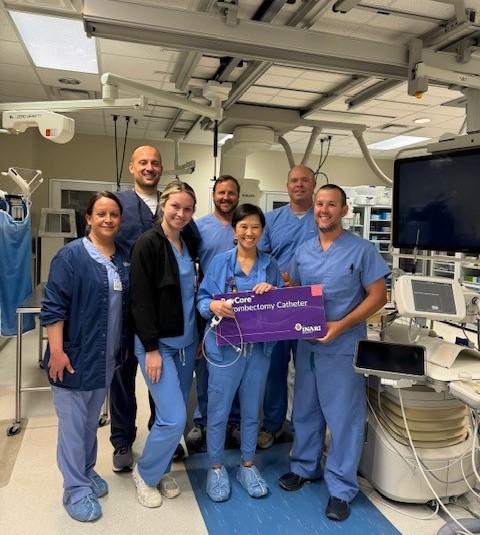 A group of seven medical professionals, including two women in black jackets and five in blue scrubs, stand in a hospital room holding a purple sign that reads, "Core Invenectomy Catheter." They are smiling and surrounded by medical equipment.