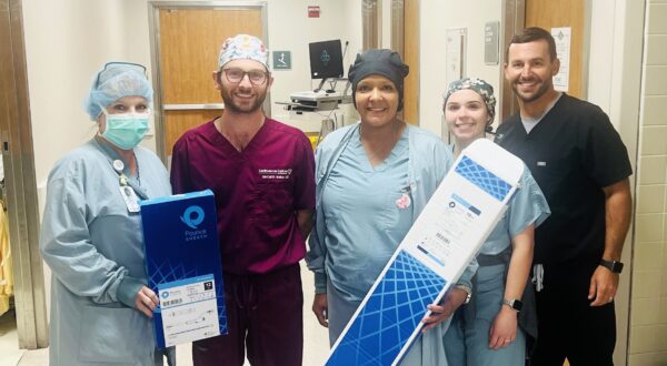 Five medical professionals in scrubs stand together in a hospital hallway, showcasing the latest venous thrombectomy system. With beaming smiles, they hold two boxed medical supplies from the Cardiovascular Institute of the South, ready to advance care for patients with venous disease.