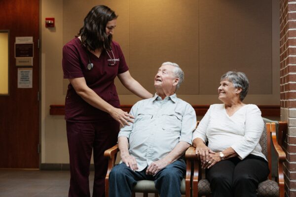 A healthcare worker in scrubs speaks with an elderly man seated next to an elderly woman on a bench in a waiting area.