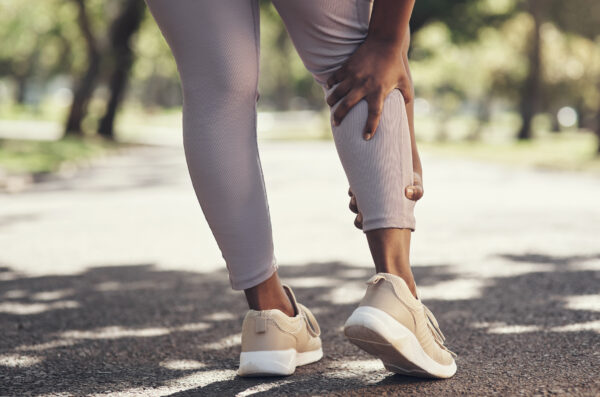 A person in beige sneakers and light gray leggings grasps their lower leg, standing on a paved pathway outdoors.