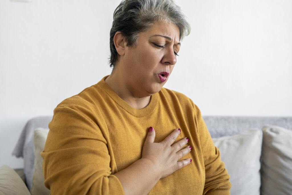 A person with short gray hair and a mustard sweater holds their chest, appearing concerned.