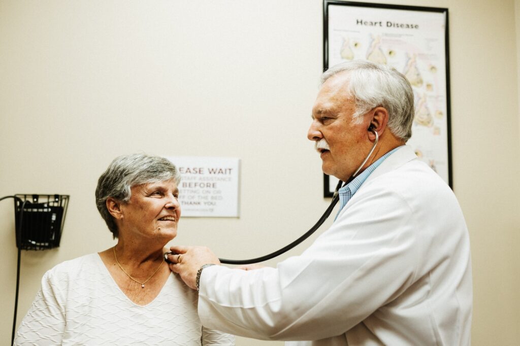 A doctor in a white coat uses a stethoscope to examine an older woman sitting in an office with a poster about heart disease on the wall.