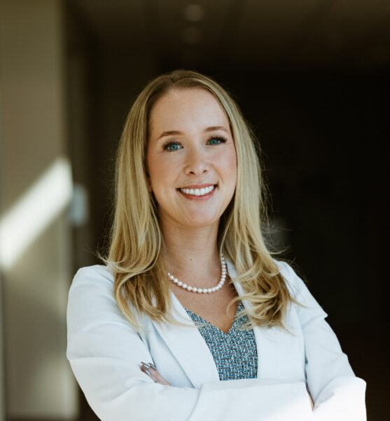 A woman with long blonde hair and a white blazer smiling, standing with arms crossed in a well-lit indoor setting.