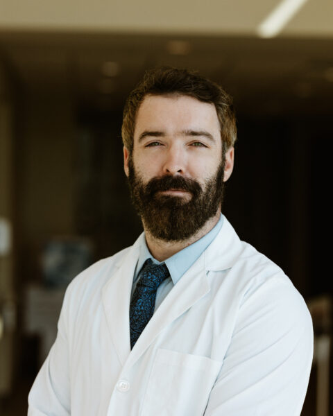 A man with a beard wearing a lab coat and blue tie stands with his arms crossed, looking directly at the camera.