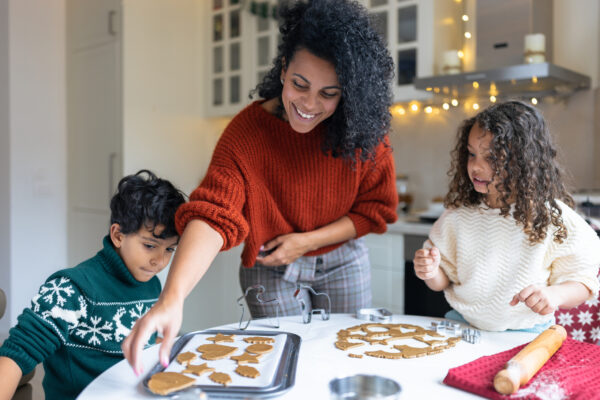 An adult and two children are making gingerbread cookies in a kitchen. The table is filled with dough, cookie cutters, and baking tools.