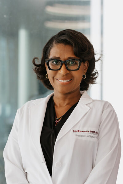 A person wearing glasses and a white lab coat stands indoors, smiling and looking at the camera.