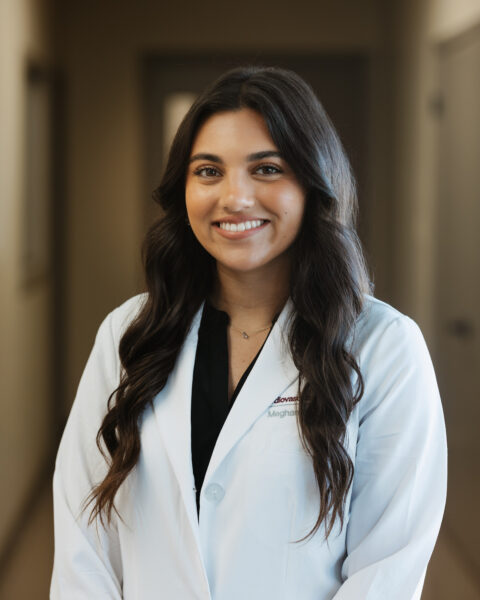 A person wearing a white lab coat smiles while standing in a hallway.