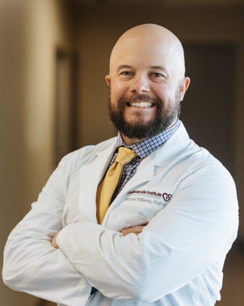 A bald man with a beard is smiling, standing with his arms crossed, wearing a white lab coat and a yellow tie.