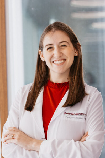 A person wearing a white lab coat with a name embroidered on it smiles while standing with arms crossed.