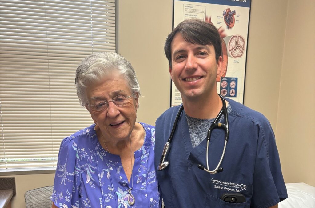 An older woman in a floral blouse stands next to a man in blue medical scrubs with a stethoscope, in an office with a heart anatomy poster.